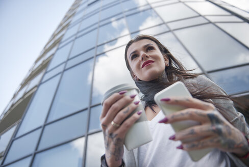 Geschäftsfrau vor Bürogebäude, hält Kaffeetasse und Smartphone, Tattoos an den Händen, Tiefblick - CUF05978