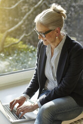 Ältere Geschäftsfrau tippt auf einem Laptop zu Hause auf der Fensterbank - CUF06025