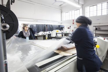 Weibliche Fabrikarbeiterinnen rollen in einer Bekleidungsfabrik Textilien auf eine Musterschneidemaschine ab - CUF06384