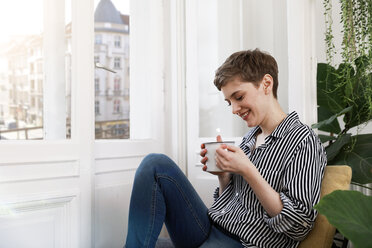 Glückliche Frau sitzt entspannt am Fenster und trinkt Kaffee - FKF02887
