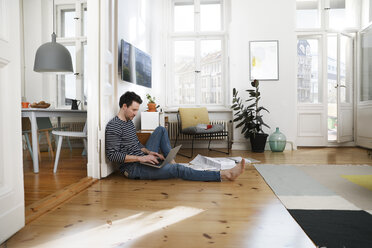 Mann sitzt auf dem Fußboden und arbeitet mit seinem Laptop von zu Hause aus - FKF02902