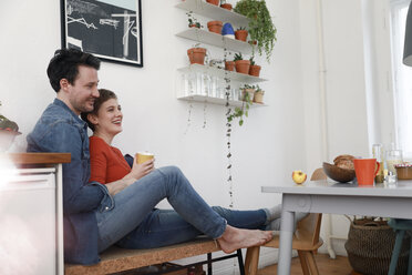 Happy couple sitting in kitchen, leaning against each other - FKF02933