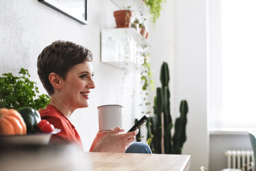 Woman sitting in kitchen, drinking coffee and checking smartphone messages - FKF02936