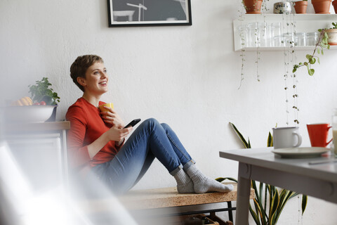 Frau sitzt in der Küche, trinkt Kaffee und prüft Smartphone-Nachrichten, lizenzfreies Stockfoto