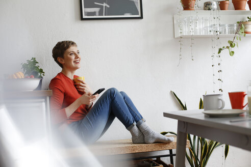 Frau sitzt in der Küche, trinkt Kaffee und prüft Smartphone-Nachrichten - FKF02939