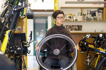 Woman in wheelchair in bicycle repair shop, holding bicycle wheel - CUF06403