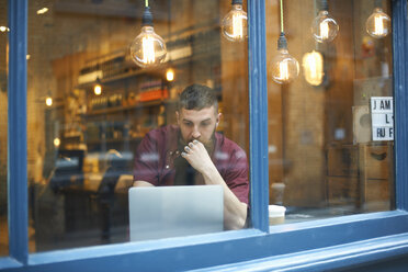 Fensteransicht eines jungen Mannes, der in einem Café einen Laptop benutzt - CUF06474