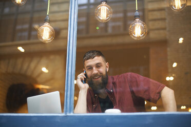 Window view of young man making smartphone call in cafe - CUF06476