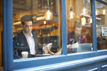 Blick aus dem Fenster auf Geschäftsleute, die in einem Café Zeitung lesen und einen Laptop benutzen - CUF06479