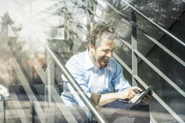 Blick aus dem Bürofenster auf einen Geschäftsmann, der auf der Treppe einen Touchscreen mit einem digitalen Tablet benutzt - CUF06514