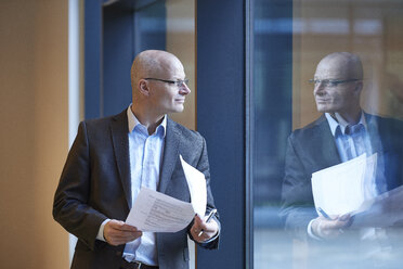 Mature businessman holding paperwork looking through office window - CUF06534