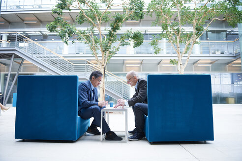 Zwei Geschäftsleute, die sich in einem Büro im Atrium in Sesseln unterhalten - CUF06542