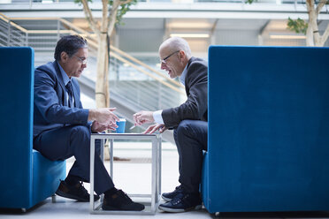 Two businessmen having discussion meeting in office atrium armchairs - CUF06543