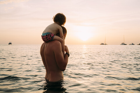 Back view of father carrying his little daughter on shoulders in the sea watching sunset stock photo