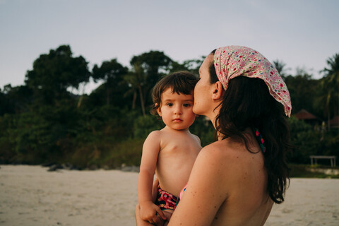 Mutter hält und küsst ihre kleine Tochter am Strand bei Sonnenuntergang, lizenzfreies Stockfoto