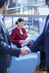 Geschäftsleute schütteln sich die Hände und schauen auf eine Geschäftsfrau, die im Atrium eines Büros Papierkram liest - CUF06626