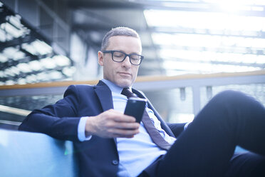 Businessman on sofa looking at smartphone in office atrium - CUF06634