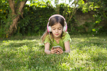 Portrait of little girl lying on meadow listening music with headphones using smartphone - LVF06984