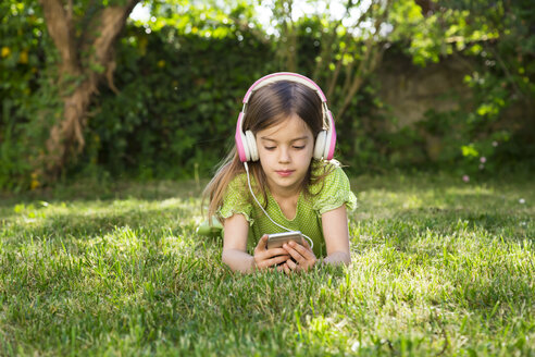 Porträt eines kleinen Mädchens, das auf einer Wiese liegt und mit Kopfhörern und Smartphone Musik hört - LVF06984