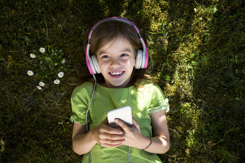 Porträt eines kleinen Mädchens, das auf einer Wiese liegt und mit Kopfhörern und Smartphone Musik hört - LVF06985