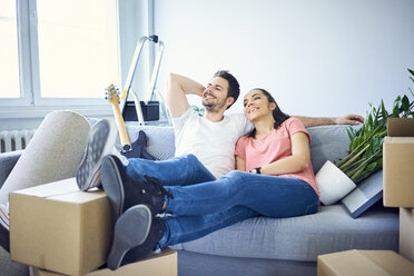 Happy couple sitting on couch surrounded by cardboard boxes - BSZF00400