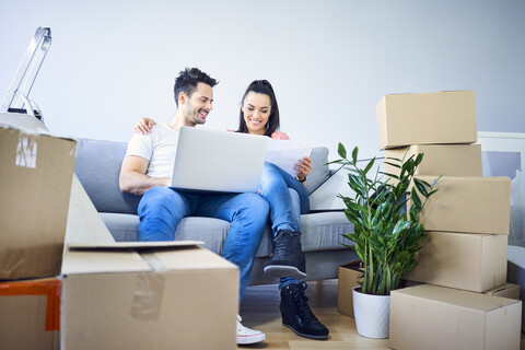 Happy couple sitting on couch surrounded by cardboard boxes using laptop stock photo