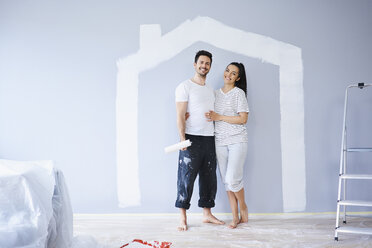 Portrait of happy couple painting in new apartment with house shape on wall - BSZF00422