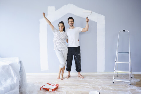 Porträt eines fröhlichen Paares in einer neuen Wohnung mit Hausform an der Wand, lizenzfreies Stockfoto