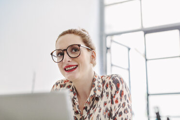 Woman in office wearing reading glasses looking away smiling - CUF06851