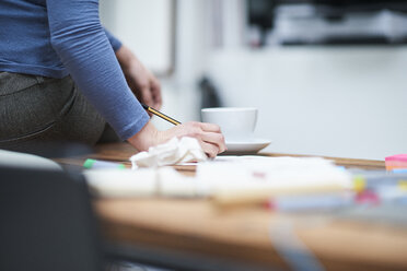 Abgeschnittene Aufnahme einer Designerin, die auf einem Schreibtisch im Designstudio sitzt und Notizen schreibt - CUF06884