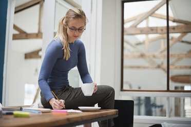 Female designer writing notes while sitting on design studio desk - CUF06885