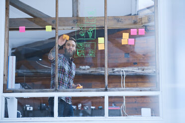 Young male designer removing adhesive note from design studio window - CUF06893