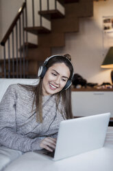 Junge Frau entspannt sich zu Hause, benutzt einen Laptop und trägt Kopfhörer - CUF07005