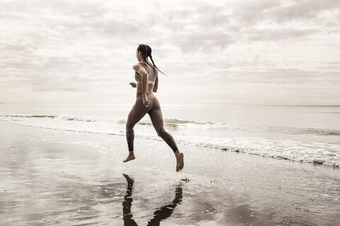 Rear view of young female runner running barefoot along water's edge at beach stock photo