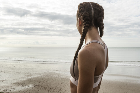 Junge Läuferin mit geflochtenen Haaren, die vom Strand aus auf das Meer schaut, lizenzfreies Stockfoto