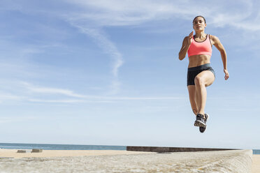 Young woman running along sea wall, mid air - CUF07188