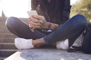 Junge Frau an der Wand sitzend, mit Smartphone, Tattoos auf der Hand, tiefer Ausschnitt - CUF07290