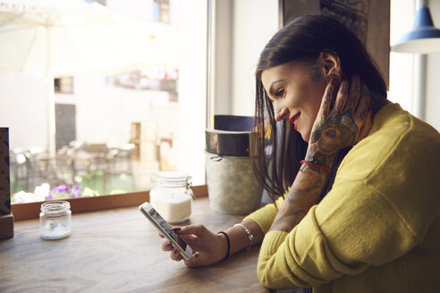 Junge Frau sitzt in einem Café und benutzt ein Smartphone, Tattoos auf Arm und Hand - CUF07292