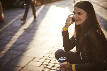 Junge Frau sitzt im Freien, hält eine Kaffeetasse, benutzt ein Smartphone, Tattoos an den Händen - CUF07293