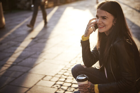 Young woman sitting outdoors, holding coffee cup, using smartphone, tattoos on hands - CUF07293