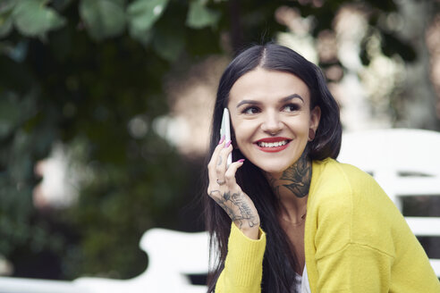 Junge Frau sitzt im Freien, benutzt ein Smartphone, lächelt, Tattoos auf Hand und Hals - CUF07295