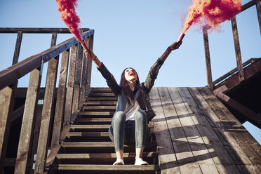 Young woman sitting on wooden steps, holding hand flares, red smoke pouring from flares, low angle view - CUF07458