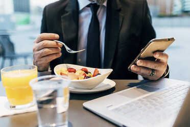 Älterer Geschäftsmann, der im Freien sitzt, frühstückt und ein Smartphone benutzt, Laptop auf dem Tisch, Mittelteil - CUF07472