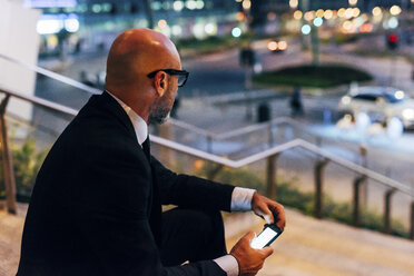 Älterer Geschäftsmann im Freien bei Nacht, auf den Stufen sitzend, Smartphone in der Hand - CUF07477