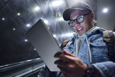 Junger Mann, der die Rolltreppe einer U-Bahn-Station hinunterfährt und auf ein digitales Tablet schaut - CUF07496