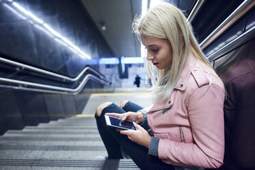 Junge Frau sitzt auf der Treppe einer U-Bahn-Station und schaut auf ihr Smartphone - CUF07498