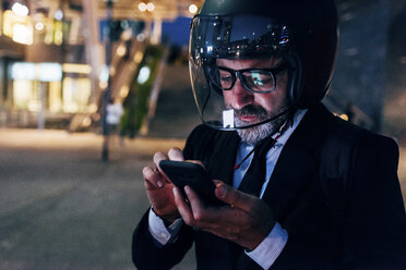 Mature businessman outdoors at night, wearing motorcycle helmet, using smartphone - CUF07504