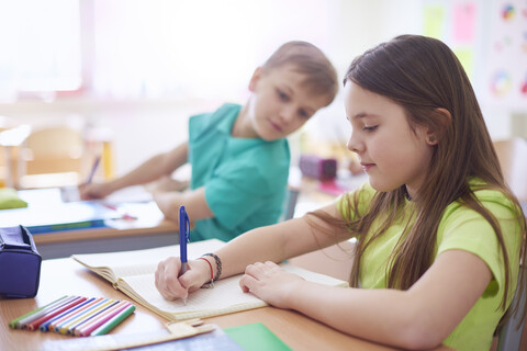 Schuljunge betrachtet Schülerin, die in der Klasse in ein Heft schreibt, lizenzfreies Stockfoto