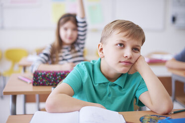 Gelangweilter Schuljunge im Unterricht mit einem Mädchen, das im Hintergrund die Hand hebt - ABIF00369