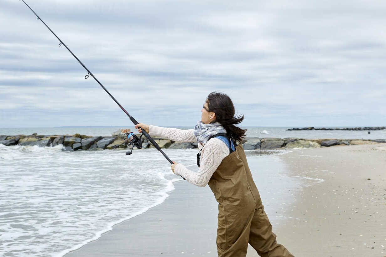Young woman in waders casting sea fishing rod from beach stock photo